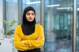 A confident woman wearing a hijab in a modern office setting, with her arms crossed. The background includes a window. photo