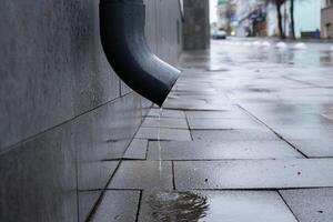 A heavy rain leaves puddles on the pavement as water drips from a pipe attached to a building in the urban area photo