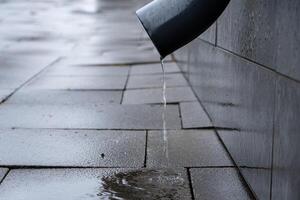 Water drips steadily from a drainpipe onto a smooth, wet pavement after recent rain, creating puddles on the ground photo