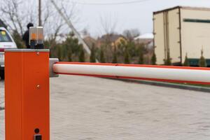 A parking lot barrier arm stands upright with a warning light on top, surrounded by vehicles and trees on a gray day photo
