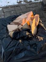 A black trash bag contains crumpled paper and empty coffee cups in a city setting with snow. The sunlight highlights the materials photo