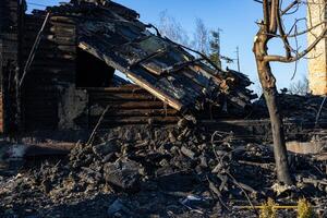 A burned-down wooden structure stands in ruin, surrounded by charred debris and ashes, with a clear sky overhead photo