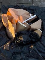 A trash bag overflowing with used coffee cups and crumpled paper waste while sunlight casts an orange hue on the litter photo