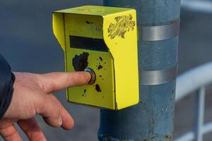 A hand is pressing a yellow pedestrian signal button located on a pole at a busy crosswalk, indicating a desire to cross the street safely photo