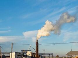 A large factory releases white smoke from its chimney against a backdrop of a clear blue sky during late afternoon hours photo