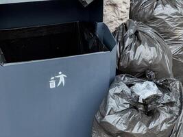 Black trash bags are piled beside a recycling bin in an urban setting, highlighting waste disposal practices in a busy area photo