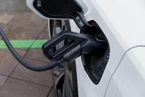 A white electric vehicle is connected to a charging station, with the charging cable securely plugged in, capturing the urban atmosphere photo