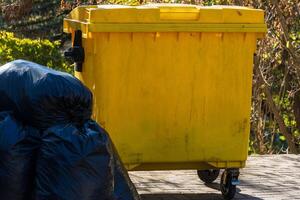 A bright yellow waste container is positioned next to several black garbage bags in a park setting during daylight hours photo