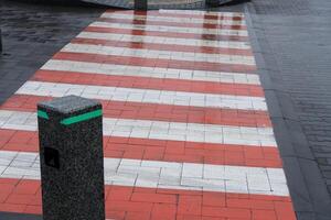 A crosswalk features vibrant red and white stripes, contrasting against wet pavement, while a decorative pole marks the area in a city photo