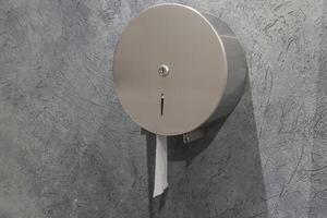 A sleek stainless steel toilet paper dispenser is attached to a textured wall, with a portion of the roll extending out for use photo