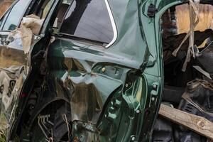 The wrecked green vehicle sits in a rural area, displaying severe dents and torn materials from an accident, surrounded by nature photo