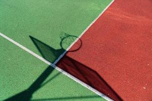 A colorful basketball court in bright sunlight shows a hoop's shadow stretching across red and green surfaces, inviting joyful games photo