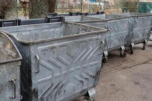 Several metal trash bins are lined up against a tree, positioned on a paved sidewalk in an urban location, under overcast skies photo