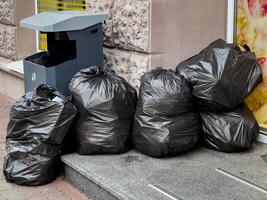 Black trash bags rest against a wall next to a recycling bin, emphasizing the need for proper waste disposal in city spaces photo