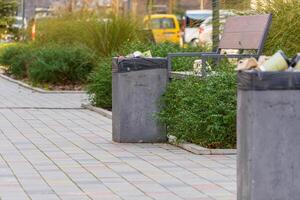 Unkempt garbage containers overflow with trash bags and recycling materials beside a pathway in an urban public area photo
