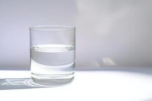 Sophisticated photograph of fresh drinking water in glass on white tabletop, with gentle shadows adding depth to minimalist scene photo