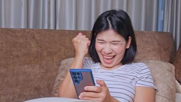 Young woman using smartphone, Smiling at her smartphone, Suddenly celebrating with joy, Smile and celebrating with excitement, Happy emotional reaction to good news, Success, at sofa living room video