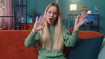 Hello. Caucasian woman smiling friendly at camera and waving hands gesturing hello, hi, greeting or goodbye, welcoming with hospitable expression at home. Teen girl sitting on sofa in living room video