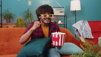 Excited indian man in 3D glasses sits on couch eating popcorn snacks and watching interesting TV serial sport game, film online social media movie content at home apartment. Guy enjoying entertainment video