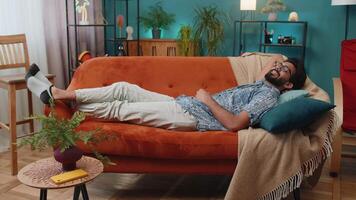 Tired indian man lying down on comfortable couch, taking a rest, daytime nap. Portrait of carefree hindu guy holding hands behind head relaxing, falling asleep at home apartment living room on sofa video