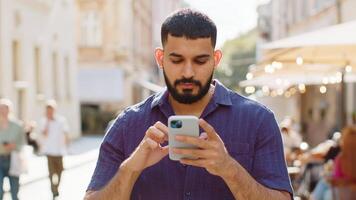 Happy smiling bearded indian man use smartphone typing text messages browsing internet social media web app working chatting online. Guy tourist looking at camera in urban city street. Town lifestyles video