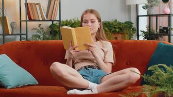 Young pretty woman relaxing reading interesting book turning pages smiling enjoying literature taking a rest on comfortable sofa. Peaceful cheerful girl at home apartment living room sitting on sofa video