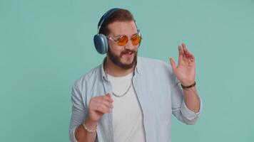 Happy lebanese man in headphones listening music and dancing disco fooling around having fun expressive gesticulating hands relaxing on party. Arabian guy isolated alone on studio blue wall background video
