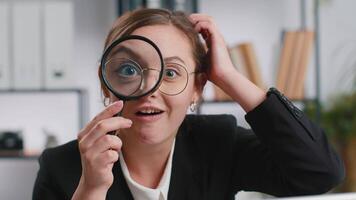 Investigator researcher scientist business woman working at office holding magnifying glass near face looking into camera with big zoomed funny eye, searching, analysing. Professional freelancer girl video
