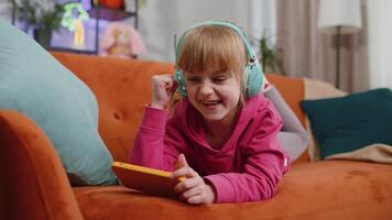 Worried funny child kid girl enthusiastically playing online racing or shooter games on smartphone. Female teen toddler using smartphone gadget app with drive simulator at home lying on couch video