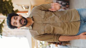 Bearded indian man smiling friendly at camera, waving hands gesturing invitation hello, hi, greeting or goodbye, welcoming with hospitable expression. Guy walking in urban city town street. Vertical video