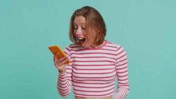 Young woman looking smartphone display sincerely rejoicing win, receiving money dollar cash banknotes, celebrating success lottery luck. Girl in crop top isolated on blue studio background. Lifestyles video
