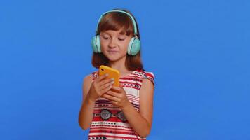 Young school girl listening music via headphones and dancing disco fooling around having fun expressive gesticulating hands. Preteen Caucasian child kid use smartphone app isolated on blue background video