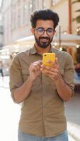 Happy smiling bearded indian man use smartphone typing text messages browsing internet social media web app working chatting online. Adult guy tourist looking at camera in urban city street. Vertical video