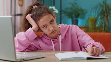 Bored tired Caucasian preteen school girl doing homework exercise online, typing on laptop, makes notes in notebook. Distance learning education. Young child kid at home workplace sitting at the table video