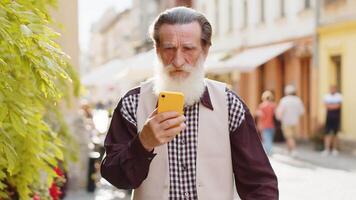 Happy smiling bearded senior old man use smartphone typing text messages browsing internet social media web app working chatting online. Mature grandfather tourist looking at camera in city street video