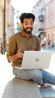 Indian man freelancer sitting in city street, looking at laptop camera, making webcam conference call with friends, family or client, enjoying distant conversation. Guy waving hello. Vertical video