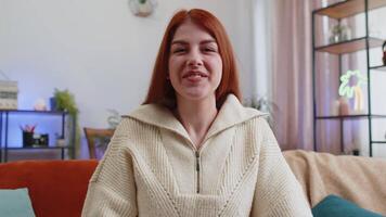 POV of young woman looking at camera, making webcam conference call with friends or family, enjoying pleasant conversation at home apartment. Redhead girl laughing, waving hello on sofa in room video