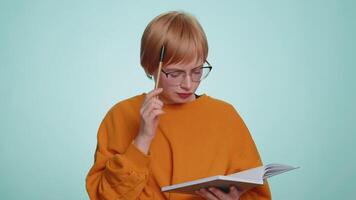 Thoughtful journalist woman making notes, writing down thoughts with pen into notepad notebook diary, to do list, good idea. Pretty short haired blonde girl isolated alone on blue studio background video