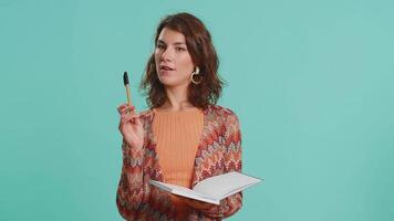 Thoughtful journalist woman making notes, writing down thoughts with pen into notepad notebook diary, to do list, good idea. Pretty brunette student girl isolated alone on blue studio background video