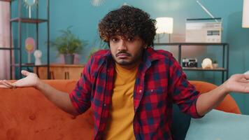 I dont know what to say. Confused indian handsome man feeling embarrassed about ambiguous question, having doubts, no idea, being clueless and uncertain at home. Hindu guy sits on sofa in living room video