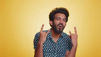 Overjoyed young indian man showing rock n roll gesture by hands, cool sign, shouting yeah with crazy expression, dancing, emotionally rejoicing in success. Hindu guy isolated on yellow background video