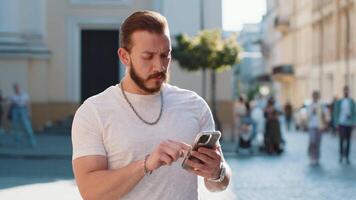 Tourist traveler bearded young man using smartphone typing text messages looking for search a way on map in mobile navigator app outdoors. Guy walking passes by urban sunshine city street background video