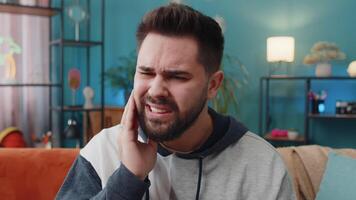 Dental problems. Young man touching cheek, closing eyes with expression of terrible suffer from painful toothache, sensitive teeth, cavities. Caucasian guy at home apartment living room on orange sofa video