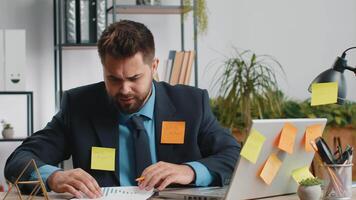 Tired exhausted Caucasian businessman working on laptop at office with many sticker task inscriptions. Freelancer man busy occupation feels sad boredom overworked multitasking. Panic attack, deadline video