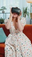 Woman using virtual reality futuristic technology VR app headset helmet to play simulation 3D 360 game, watching film movie at modern home apartment. Girl in goggles sitting on couch. Vertical video