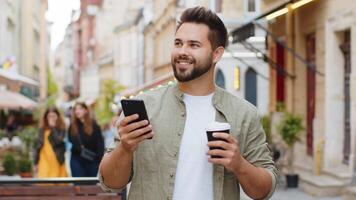 Happy smiling man use smartphone typing text messages browsing internet social media web app working chatting online enjoying morning to-go coffee hot drink outdoor. Guy tourist walking in city street video