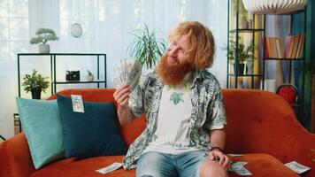 Successful happy rich Caucasian redhead man counting money cheering up with high profits, lottery game win, satisfied of wealth income earnings salary, on home room couch full of dollar cash banknotes video