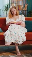 Happy red hair woman relaxing reading interesting book, turning pages smiling enjoying literature, taking a rest on comfortable couch. Redhead girl at home apartment living room on sofa. Vertical video