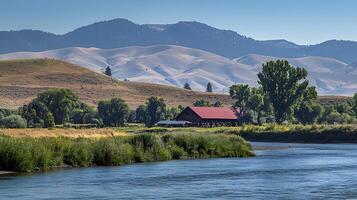 Serene Montana Barn by the River with Mountain Views. A picturesque landscape photograph showcasing a rustic barn nestled beside a calm river, set against the backdrop of majestic mountains. photo