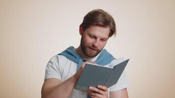 Thoughtful journalist caucasian man making notes, writing down thoughts with pen into notepad notebook diary, to do list, good idea. Handsome bearded guy isolated alone on beige studio background video